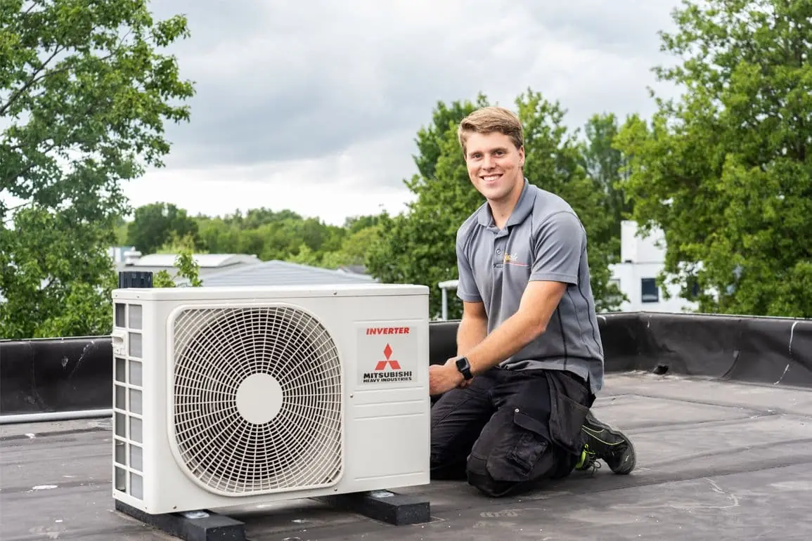 Monteur knielt glimlachend naast een Mitsubishi buitenunit van een airconditioning op een plat dak.