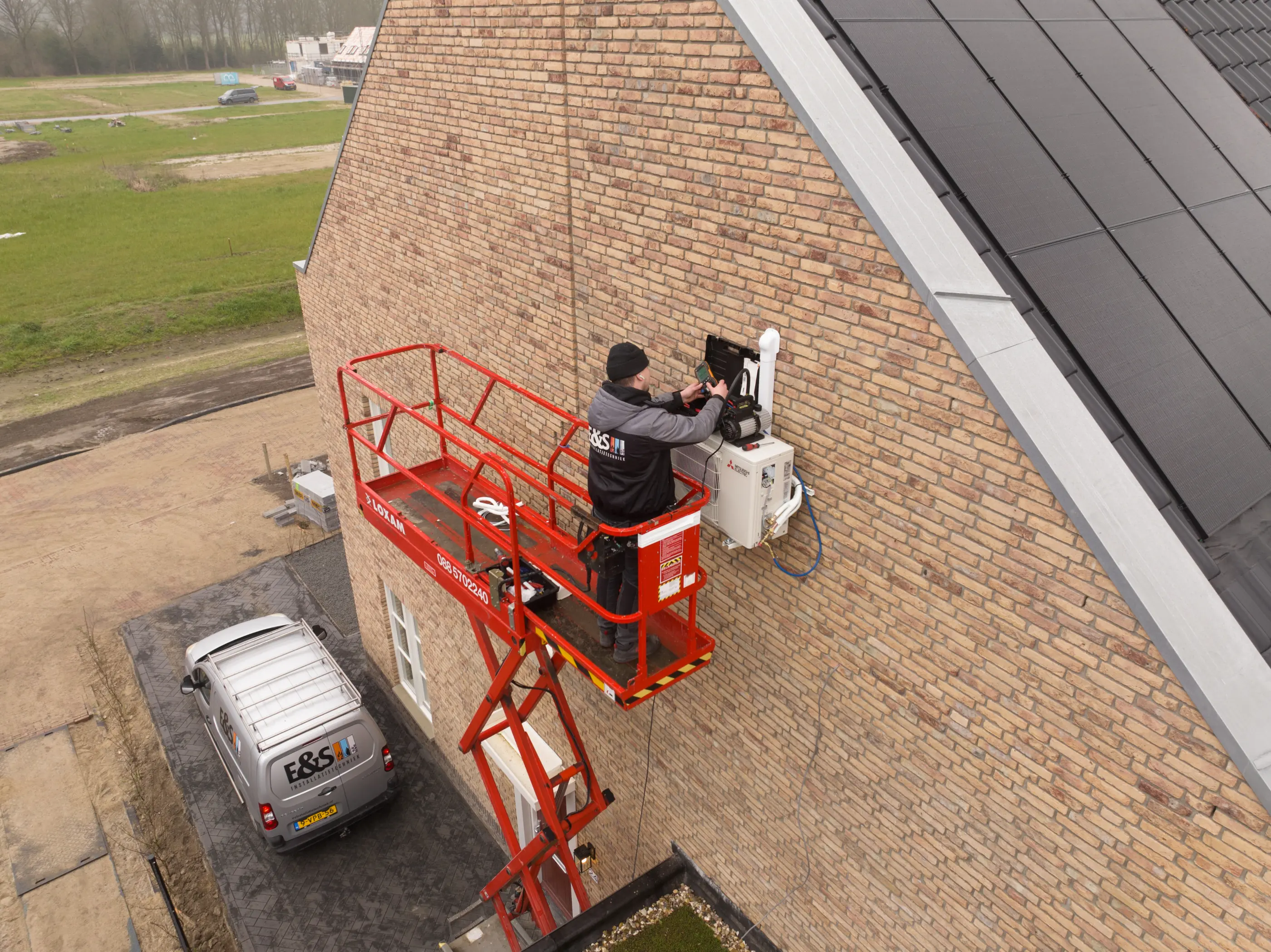Monteur op hoogwerker installeert een airconditioningunit aan de gevel van een gebouw met zonnepanelen op het dak.